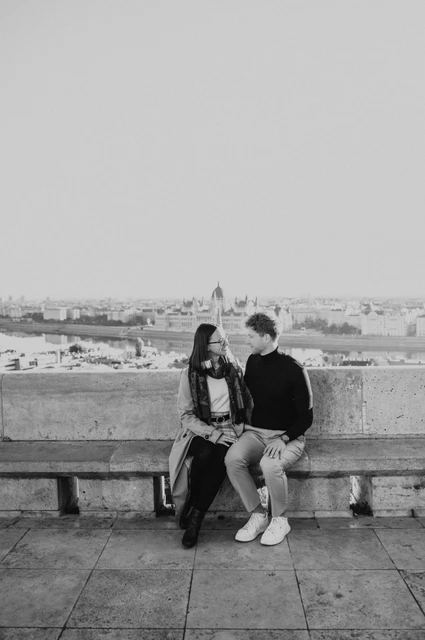 Budapest couple photoshoot: a bench portrait overlooking Parliament in black and white Black-and-white photograph of the couple sitting together on a stone bench with Parliament across the river behind them during a couple photoshoot in Budapest.