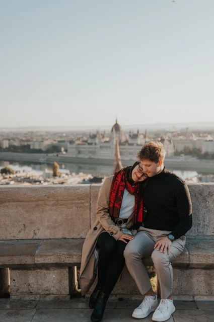 Budapest couple photoshoot: a close moment on the stone bench The man leaning towards his partner on a stone bench with Parliament behind them during a couple photoshoot in Budapest.