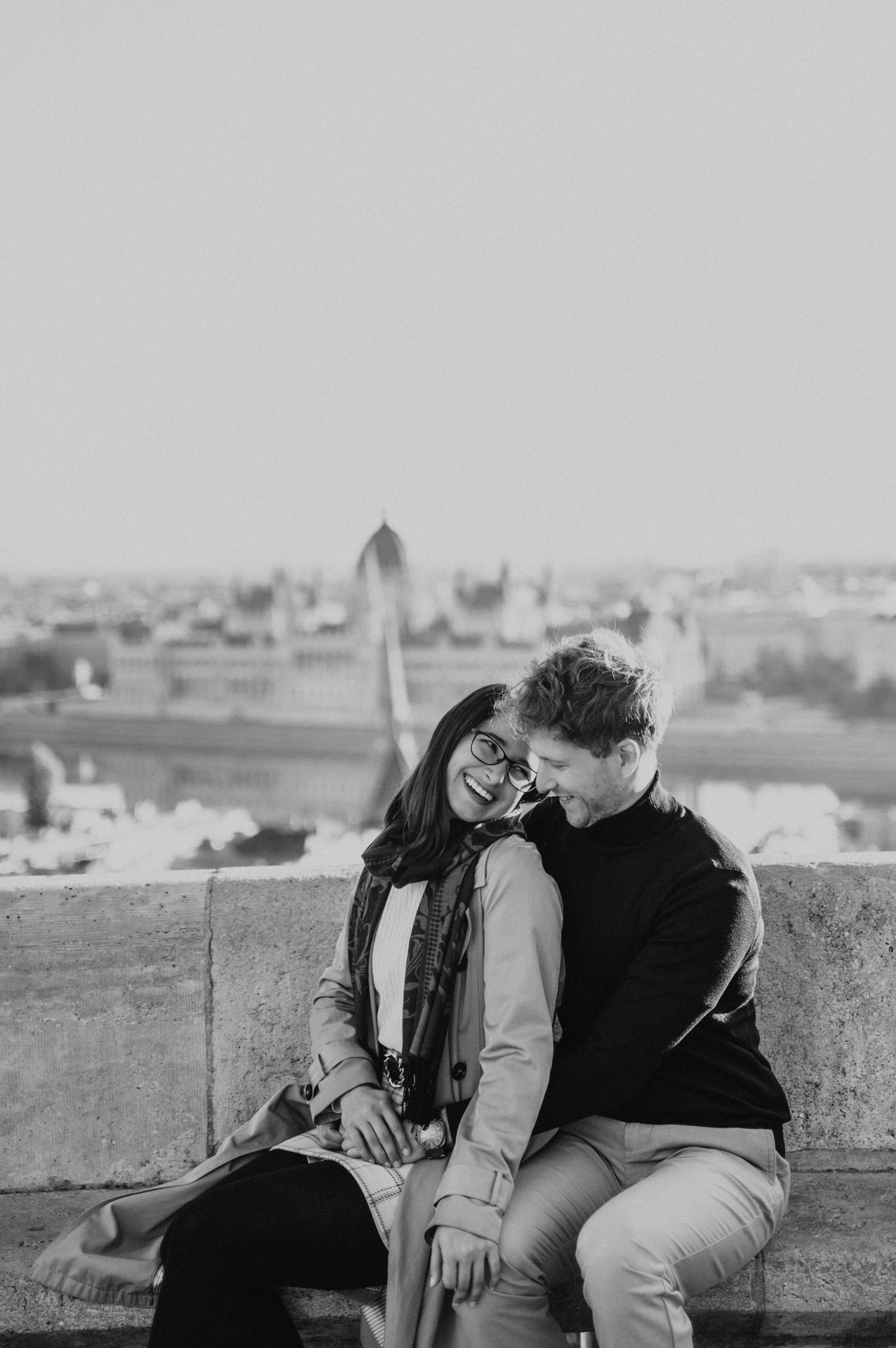 Black-and-white photograph of the couple sitting close together on a stone bench overlooking the city from Fisherman’s Bastion during a couple photoshoot in Budapest.