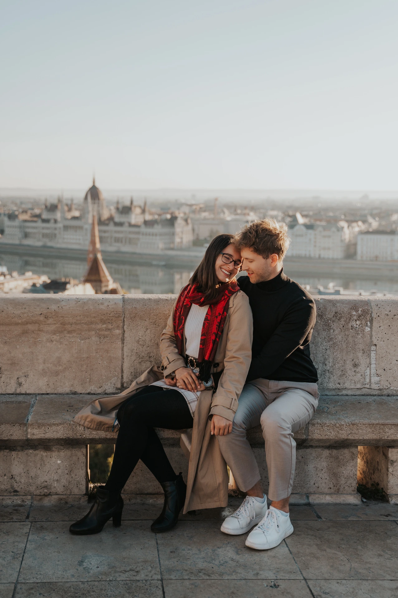 The couple leaning their heads together on a stone bench with Parliament in the background during a couple photoshoot in Budapest.