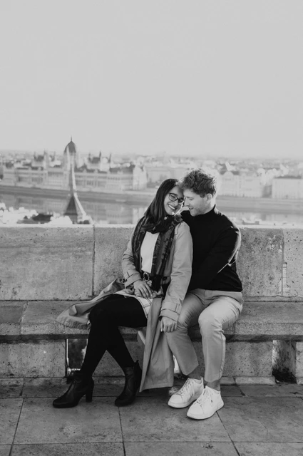 Budapest couple photoshoot: a shared portrait on the Bastion bench in black and white Black-and-white photograph of the couple sitting close and looking towards the camera on a bench at Fisherman’s Bastion during a couple photoshoot in Budapest.