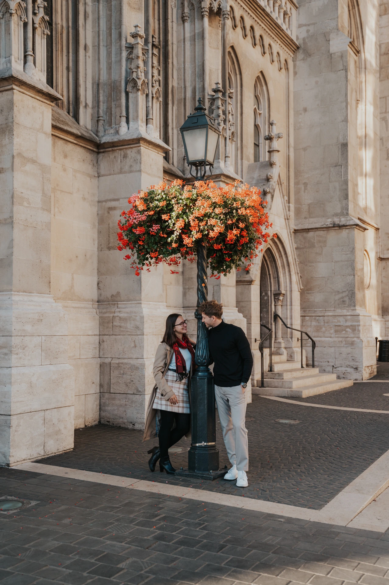 The couple standing beside a basket of orange flowers against Matthias Church’s pale stone façade during a couple photoshoot in Budapest.