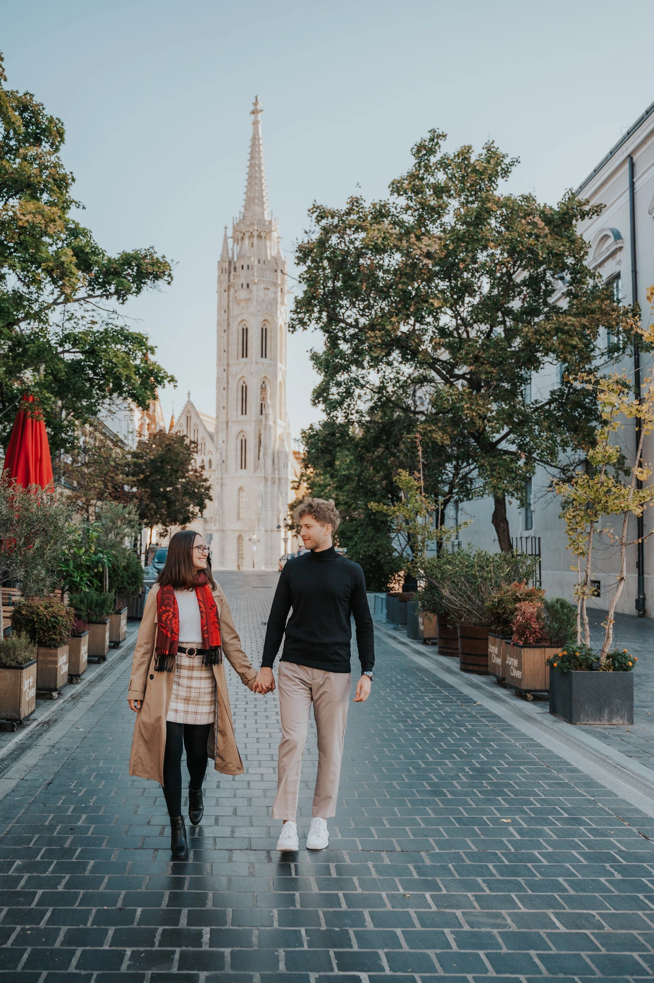 The couple walking hand in hand along a tree-lined cobbled lane with Matthias Church’s spire behind them during a couple photoshoot in Budapest.