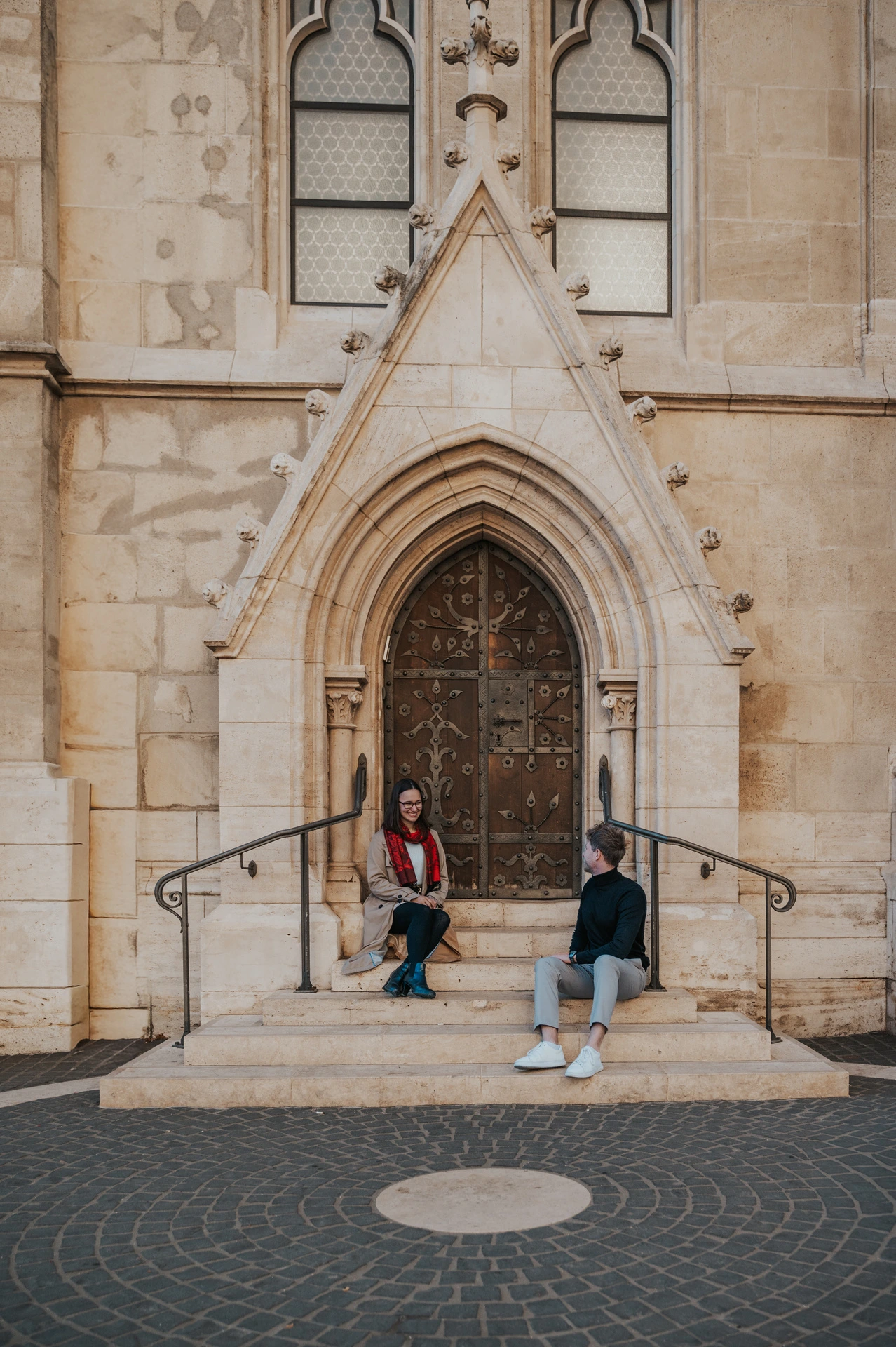 The couple sitting apart on the broad steps beneath Matthias Church’s pointed doorway during a couple photoshoot in Budapest.