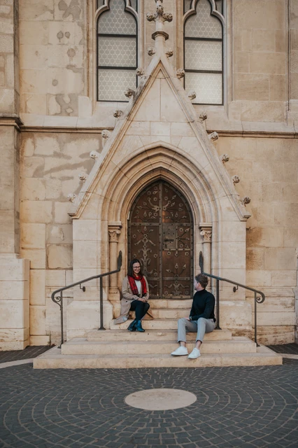 Budapest couple photoshoot: a wide view of the church steps The couple sitting apart on the broad steps beneath Matthias Church’s pointed doorway during a couple photoshoot in Budapest.