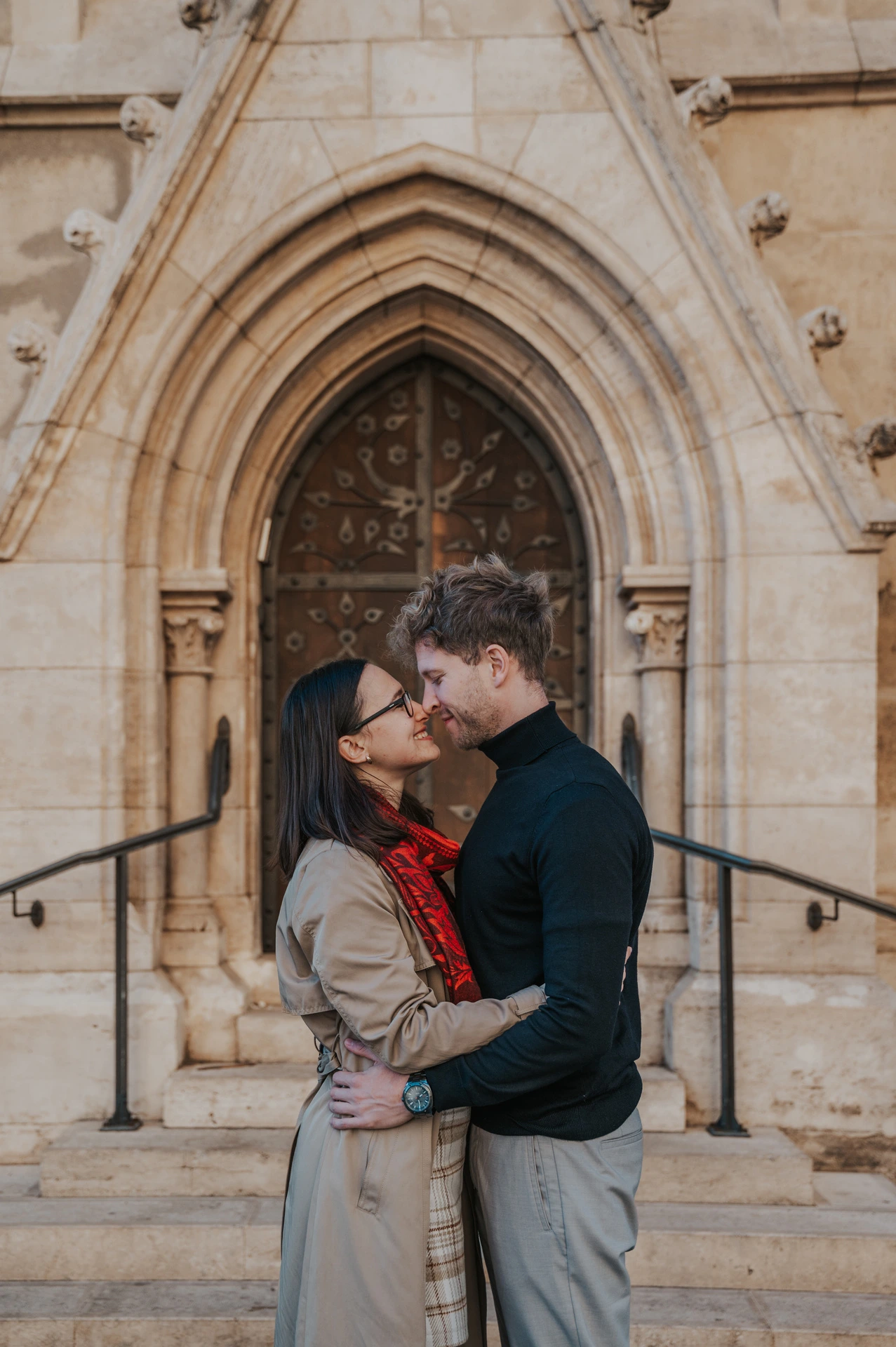 The couple embracing and leaning close outside the pointed wooden doorway at Matthias Church during a couple photoshoot in Budapest.