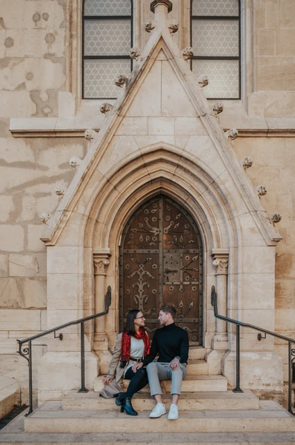 Budapest couple photoshoot: the pointed doorway of Matthias Church A wide portrait of the couple sitting on the steps below a pointed wooden doorway at Matthias Church during a couple photoshoot in Budapest.