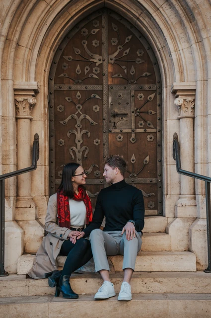 The couple sitting together on the steps in front of a carved wooden door at Matthias Church during a couple photoshoot in Budapest.