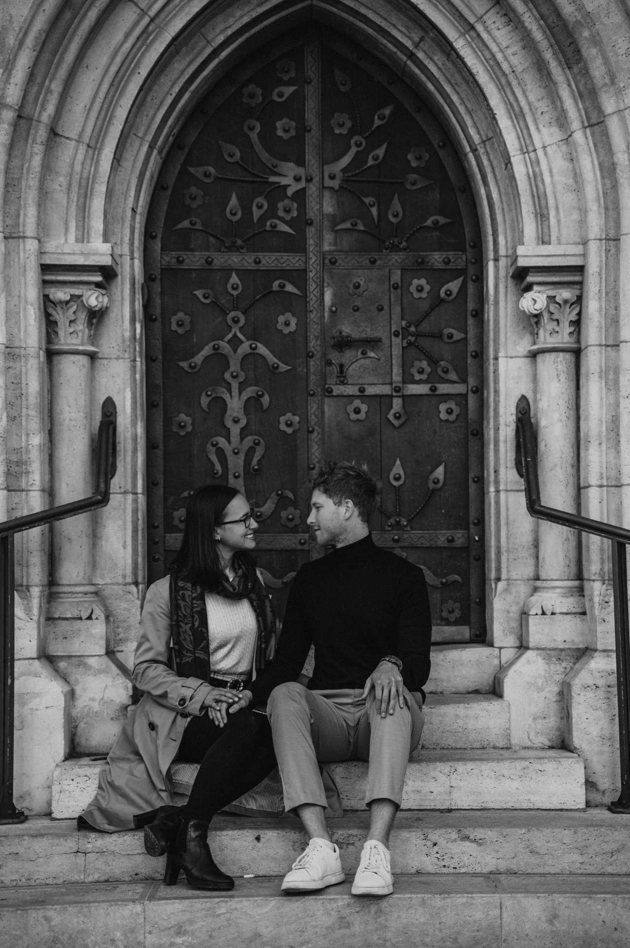 Black-and-white photograph of the couple sitting on stone steps beneath the pointed arch of a carved door at Matthias Church during a couple photoshoot in Budapest.