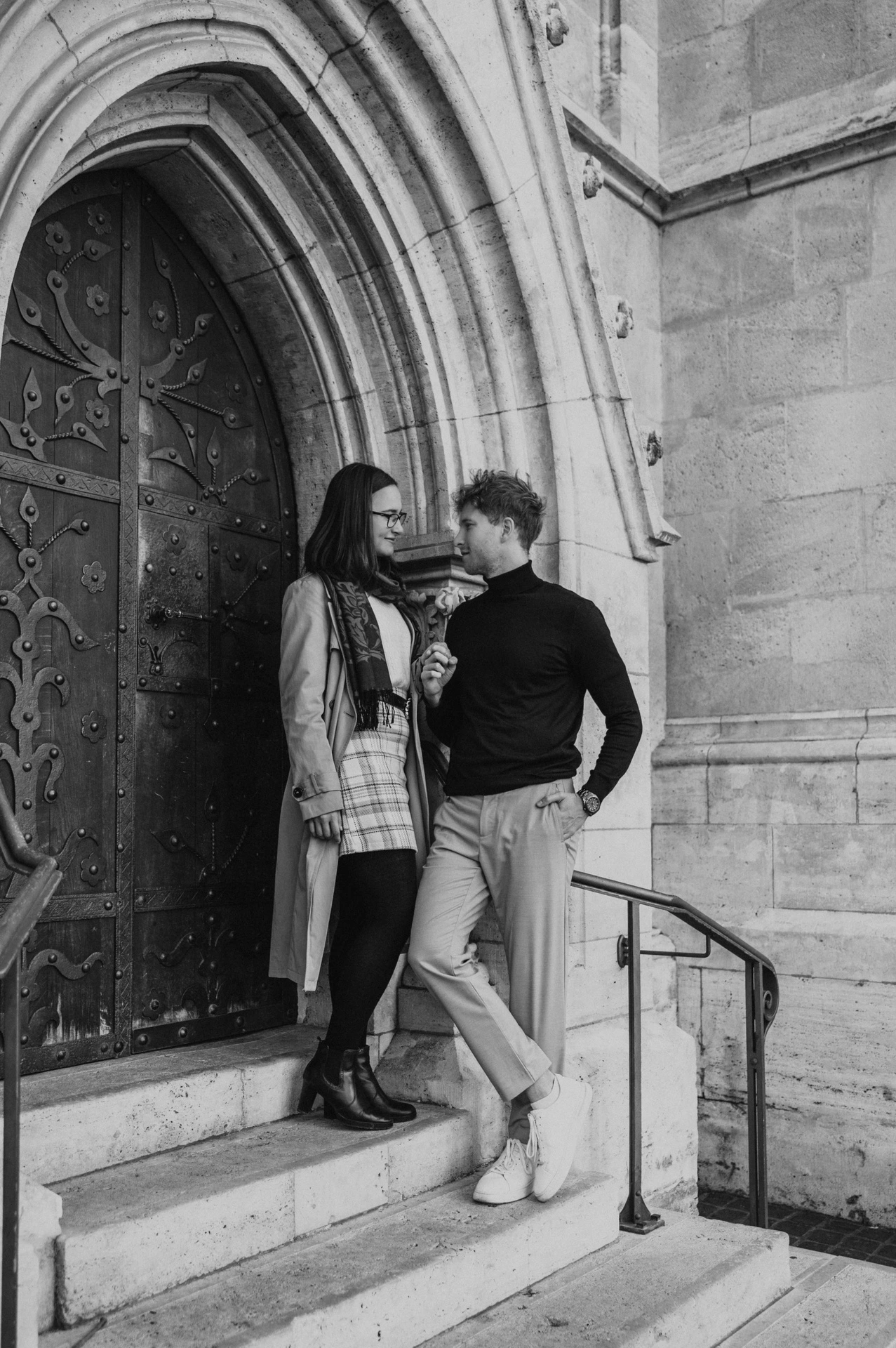 Black-and-white photograph of the couple standing close on the steps outside Matthias Church’s carved wooden door during a couple photoshoot in Budapest.