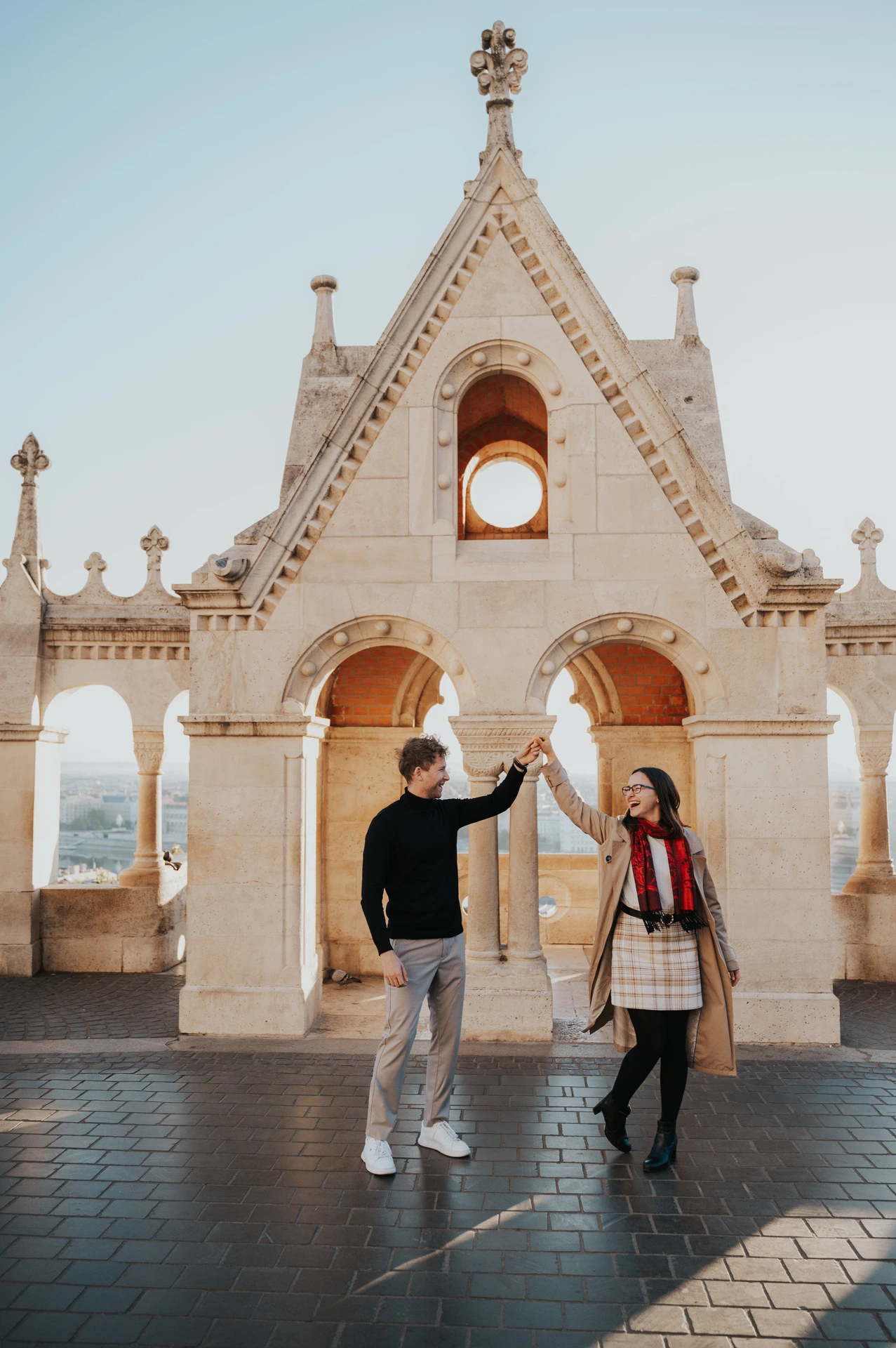The man raising his partner’s hand beneath Fisherman’s Bastion’s gabled stone arch during a couple photoshoot in Budapest.