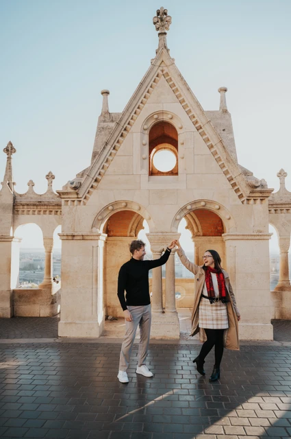 The man raising his partner’s hand beneath Fisherman’s Bastion’s gabled stone arch during a couple photoshoot in Budapest.