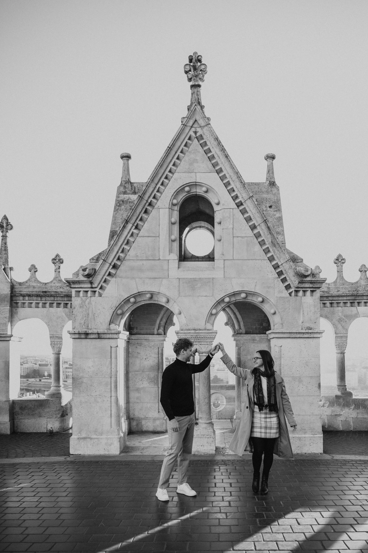 Black-and-white photograph of the couple holding hands as they turn beneath Fisherman’s Bastion’s gabled arch during a couple photoshoot in Budapest.