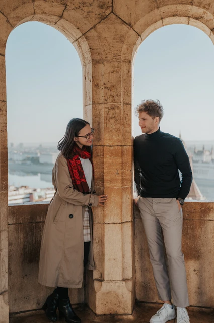 Budapest couple photoshoot: inside the Bastion’s stone arcade The couple looking towards each other from either side of a stone column inside Fisherman’s Bastion’s arcade during a couple photoshoot in Budapest.