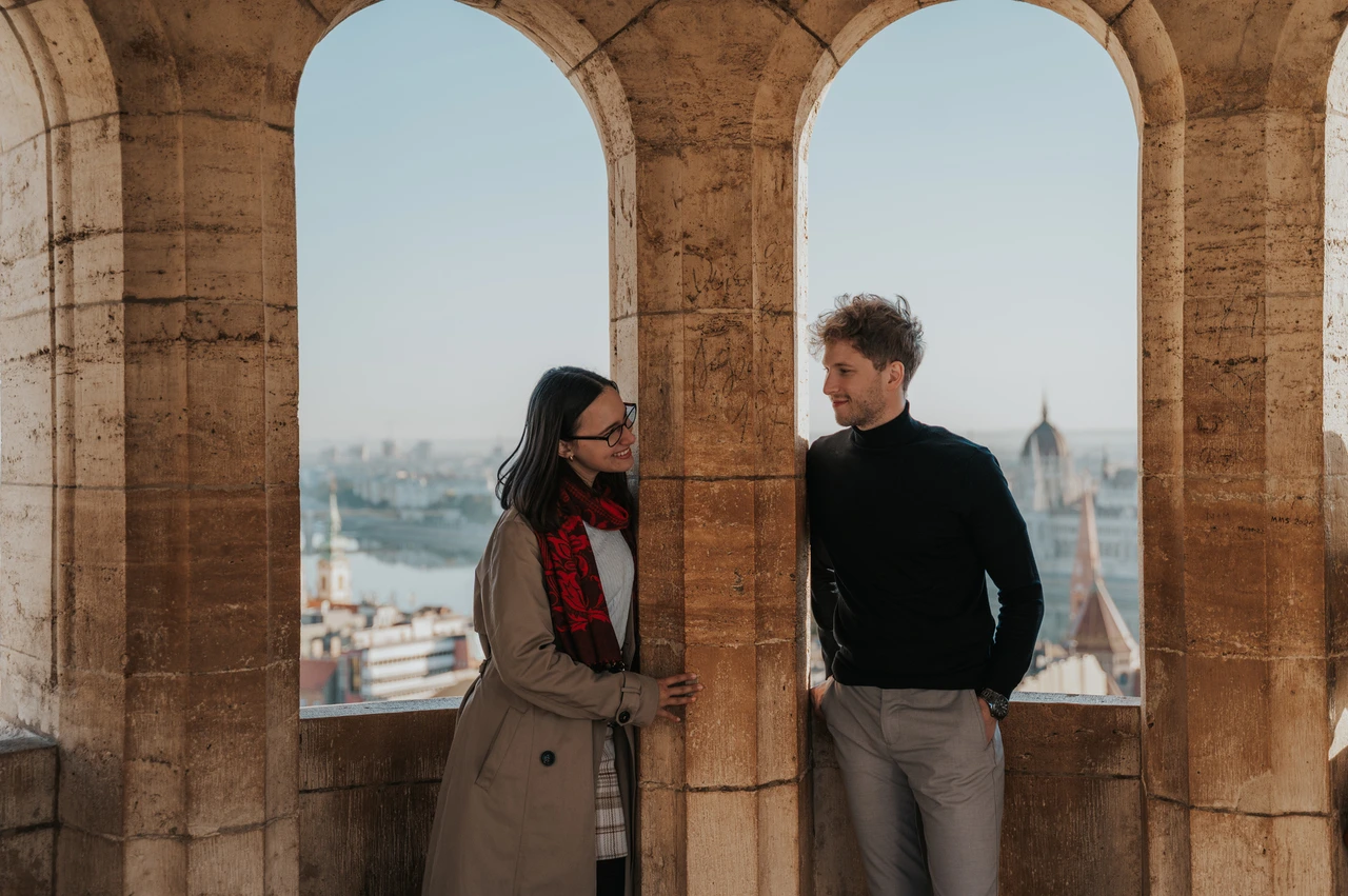 Budapest couple photoshoot: arched city views from the stone arcade A landscape portrait of the couple on either side of a stone column with arched city views at Fisherman’s Bastion during a couple photoshoot in Budapest.