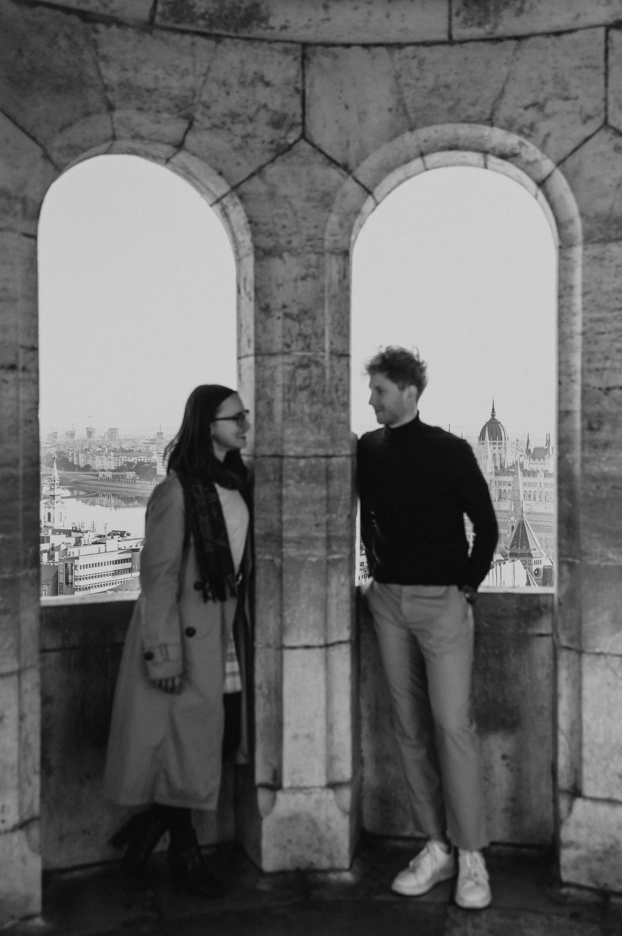 Black-and-white photograph of the couple standing beside a stone column between arched windows at Fisherman’s Bastion during a couple photoshoot in Budapest.