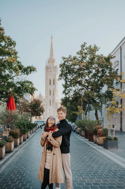Budapest couple photoshoot: an embrace on the cobbled lane The man embracing his partner from behind on a cobbled lane with Matthias Church’s spire in the background during a couple photoshoot in Budapest.