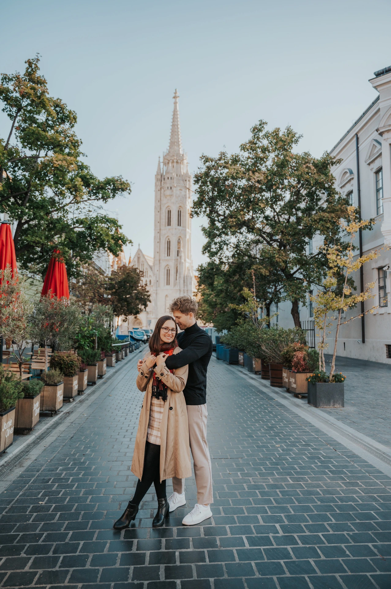 A full-length portrait of the couple embracing on a cobbled lane lined with potted plants near Matthias Church during a couple photoshoot in Budapest.