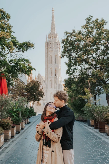 The man kissing his smiling partner near her temple on a tree-lined lane with Matthias Church beyond during a couple photoshoot in Budapest.