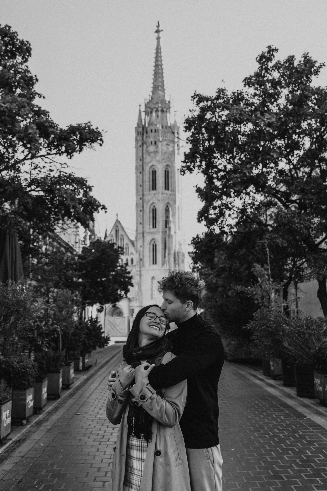 Black-and-white photograph of the couple standing close together on a tree-lined lane near Matthias Church during a couple photoshoot in Budapest.