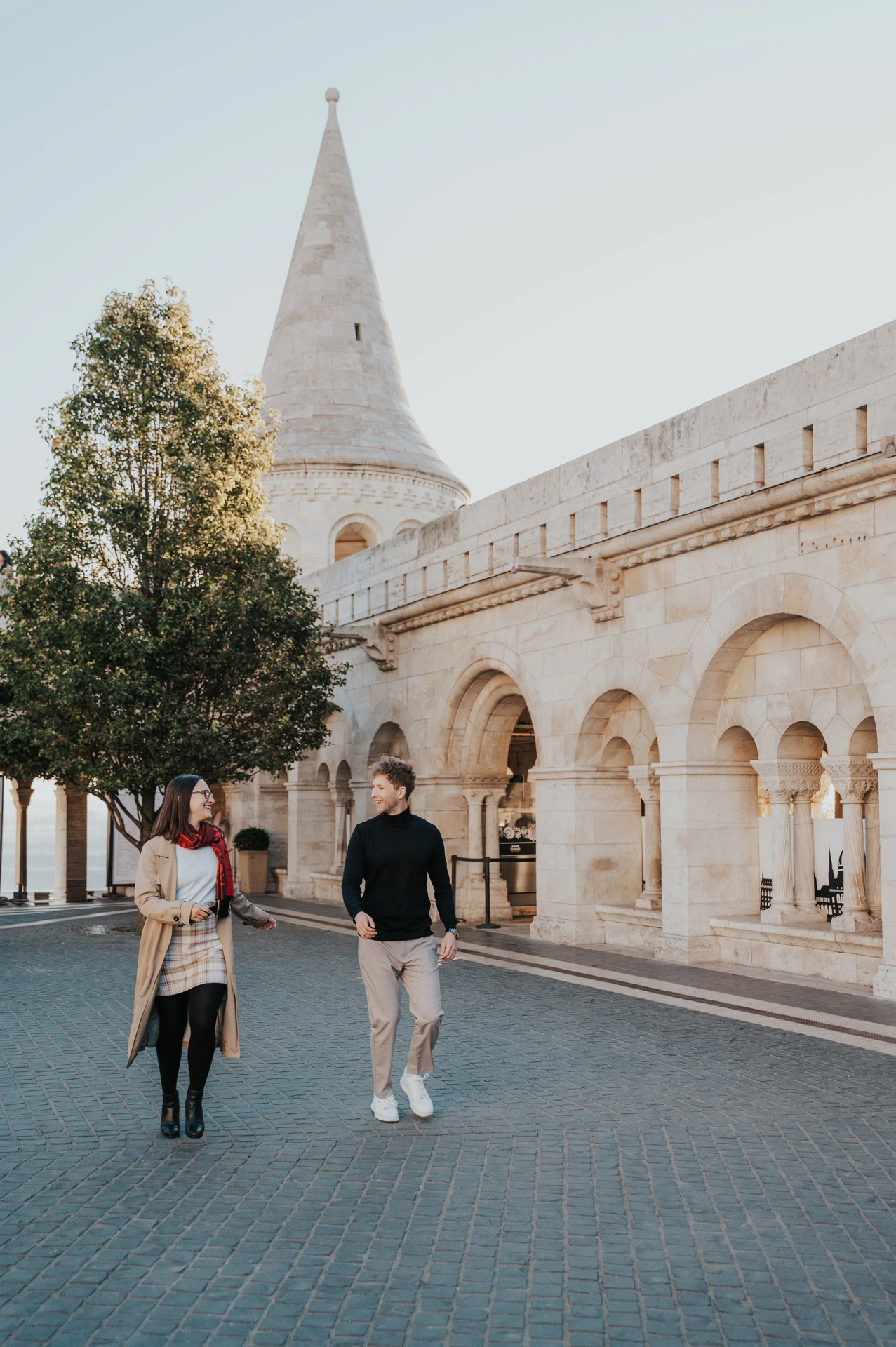 The couple walking across the square beside Fisherman’s Bastion’s pale stone arcade and autumn trees during a couple photoshoot in Budapest.