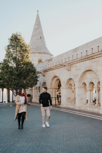 Budapest couple photoshoot: a walk beside the Bastion arcade The couple walking across the square beside Fisherman’s Bastion’s pale stone arcade and autumn trees during a couple photoshoot in Budapest.