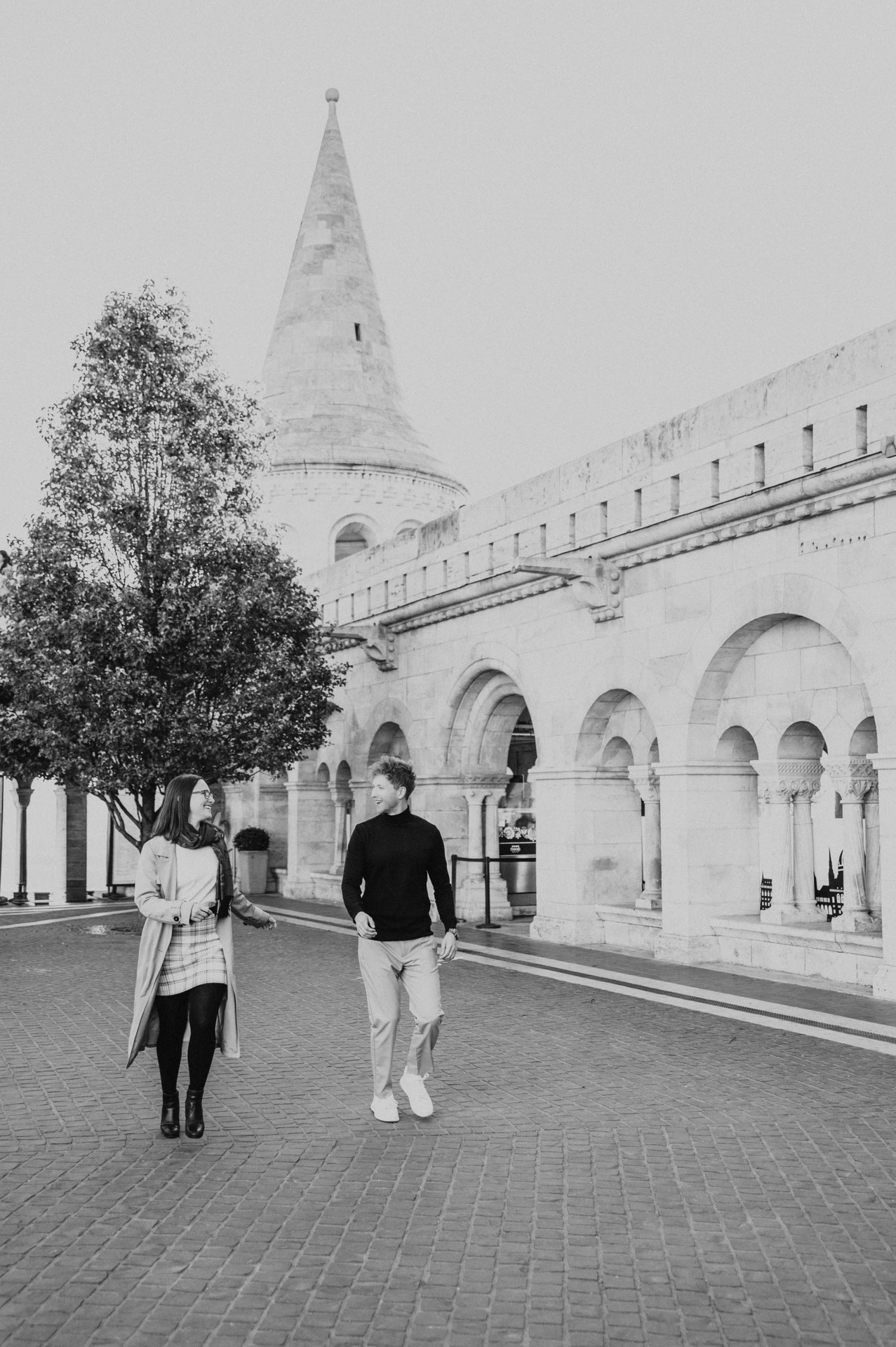 Black-and-white photograph of the couple walking side by side past Fisherman’s Bastion’s stone arcade during a couple photoshoot in Budapest.