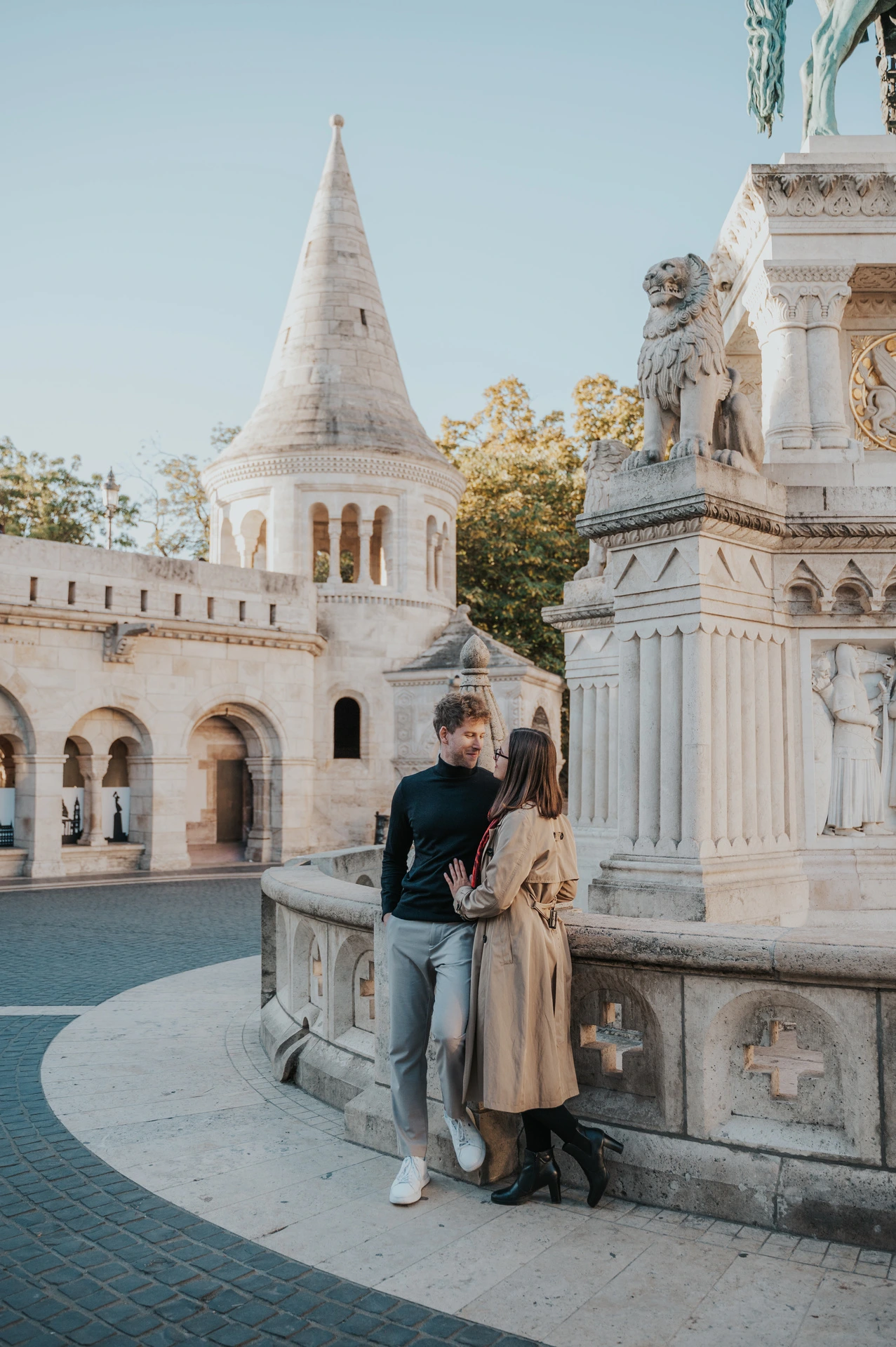 The couple beside a carved stone monument with Fisherman’s Bastion’s tower and arcade behind them during a couple photoshoot in Budapest.