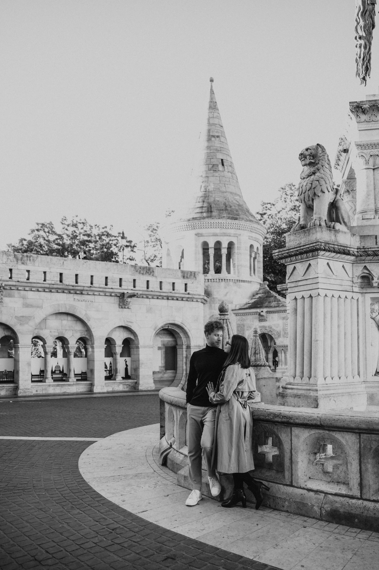 Black-and-white photograph of the couple beside a carved monument with Fisherman’s Bastion’s pointed tower behind them during a couple photoshoot in Budapest.