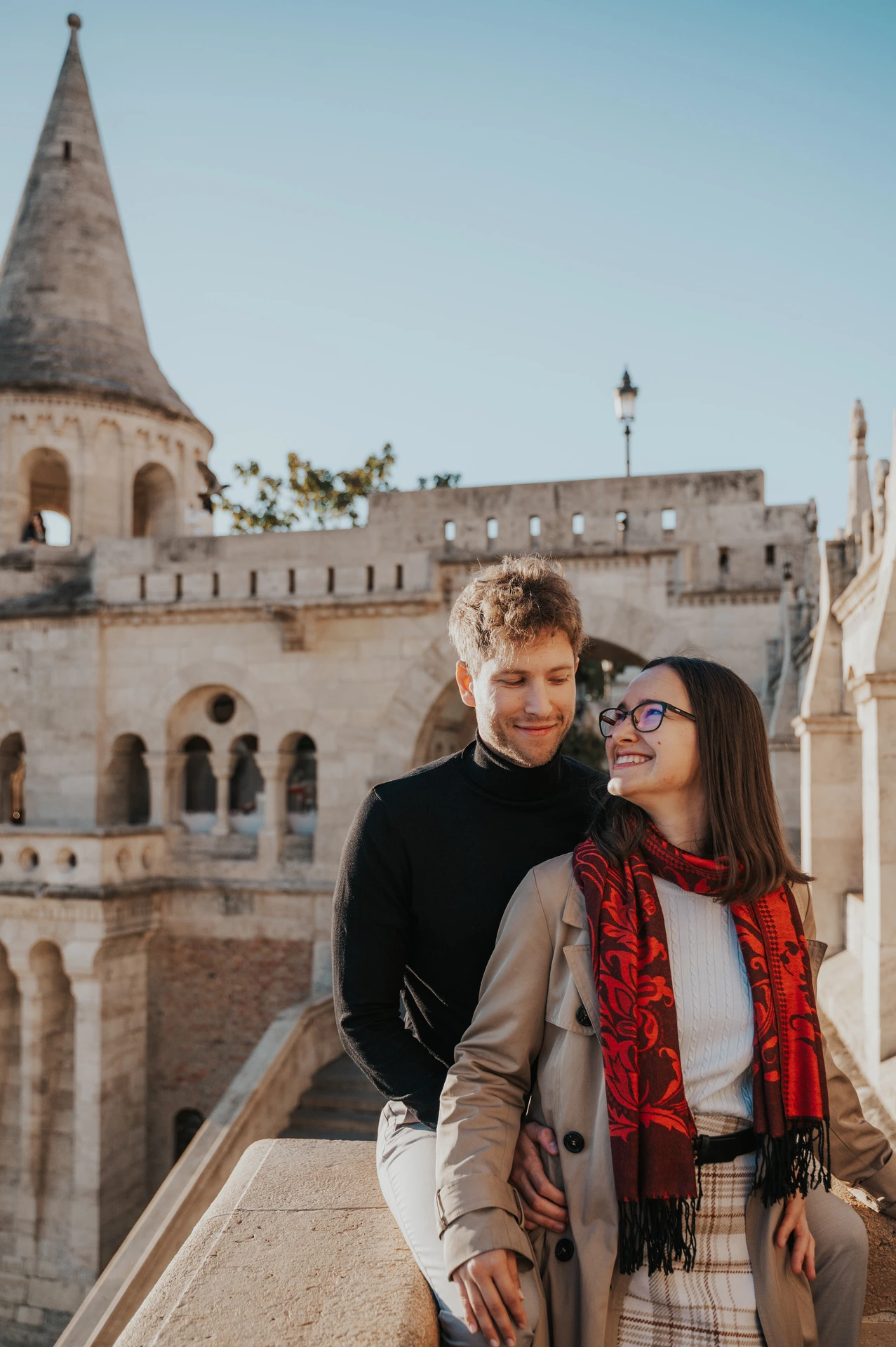 The smiling couple standing close beside a stone balustrade with Fisherman’s Bastion’s tower behind them during a couple photoshoot in Budapest.
