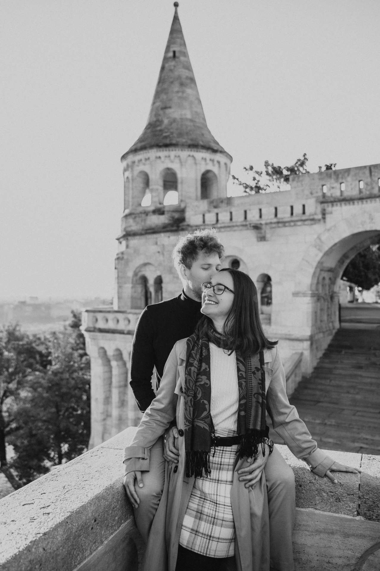 Black-and-white photograph of the couple standing close with a pointed tower at Fisherman’s Bastion behind them during a couple photoshoot in Budapest.