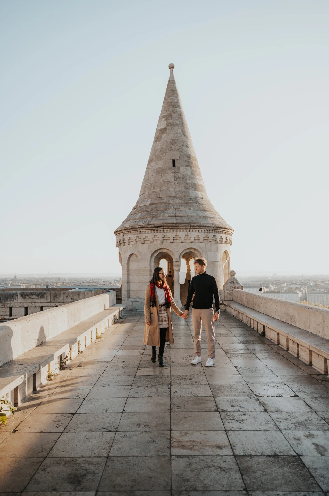 The couple walking along a stone terrace towards a pointed tower at Fisherman’s Bastion during a couple photoshoot in Budapest.