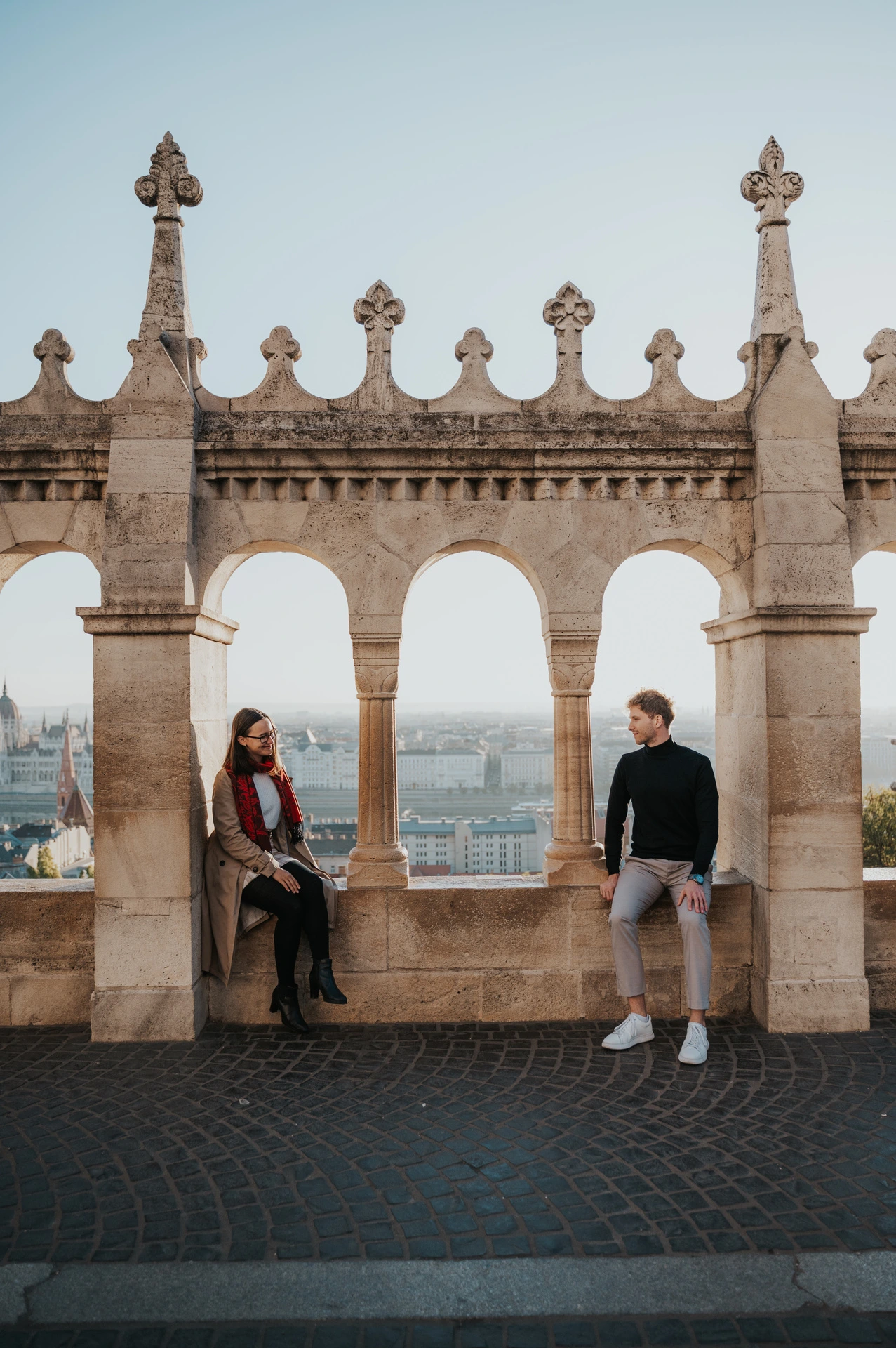 The couple on opposite sides of a stone archway at Fisherman’s Bastion, with Parliament across the Danube during a couple photoshoot in Budapest.