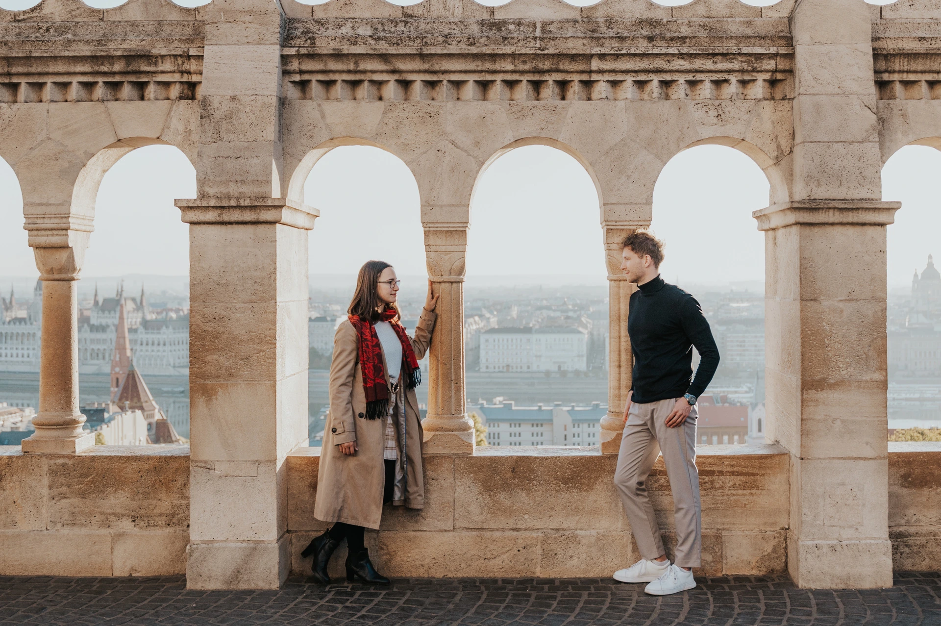 Budapest couple photoshoot: a wide Parliament view through the arches A wide view of the couple on opposite sides of a stone arch at Fisherman’s Bastion with Parliament beyond during a couple photoshoot in Budapest.