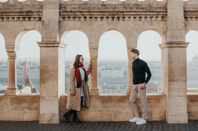 A wide view of the couple on opposite sides of a stone arch at Fisherman’s Bastion with Parliament beyond during a couple photoshoot in Budapest.
