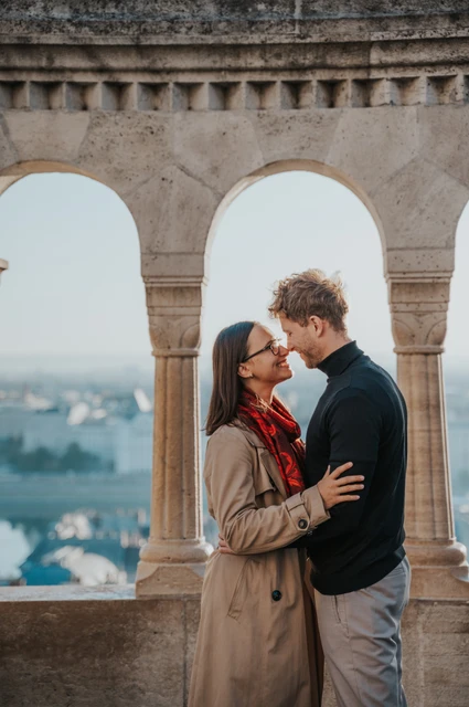 Budapest couple photoshoot: a red scarf beneath the stone arches The smiling couple leaning towards each other beneath Fisherman’s Bastion’s stone arches, with the woman’s red scarf in view during a couple photoshoot in Budapest.