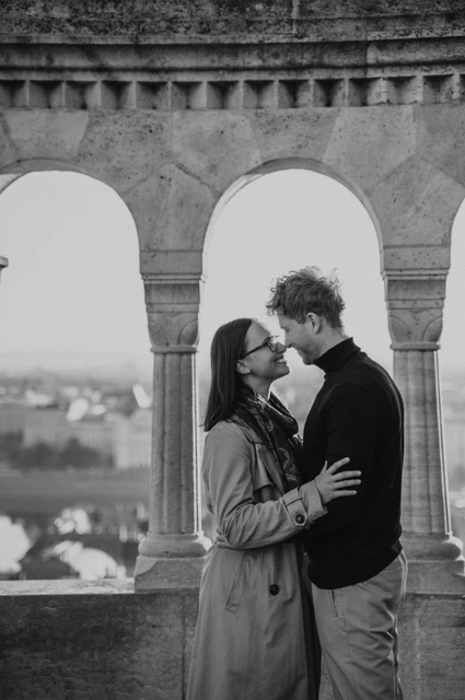 Budapest couple photoshoot: a close portrait in the stone arcade in black and white Black-and-white photograph of the couple leaning close beneath Fisherman’s Bastion’s stone arcade with the city in the background during a couple photoshoot in Budapest.