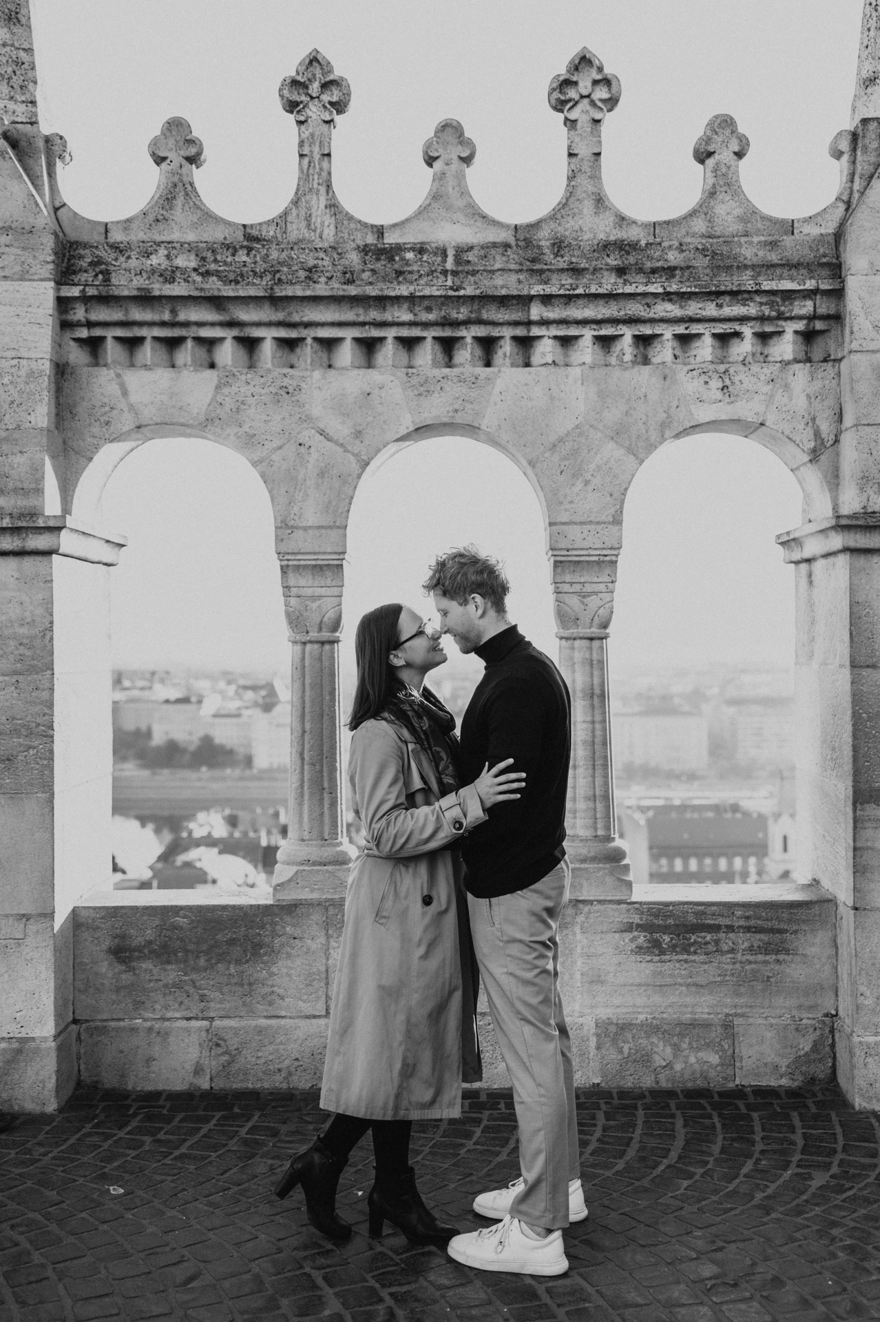 Black-and-white photograph of the couple standing close beneath the carved stone arches at Fisherman’s Bastion during a couple photoshoot in Budapest.