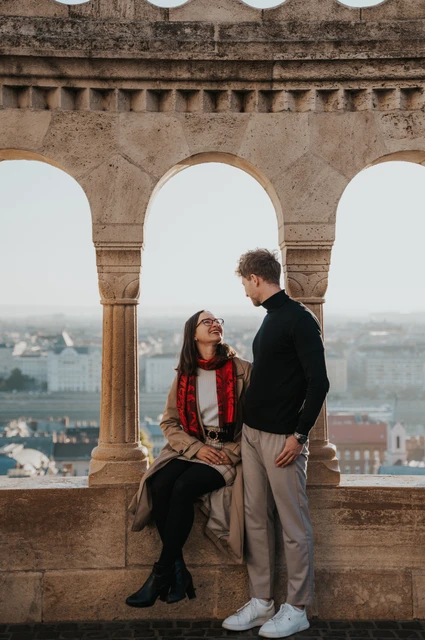 Budapest couple photoshoot: an arched window overlooking the city The woman sitting in an arched opening beside her standing partner at Fisherman’s Bastion, with the city beyond during a couple photoshoot in Budapest.