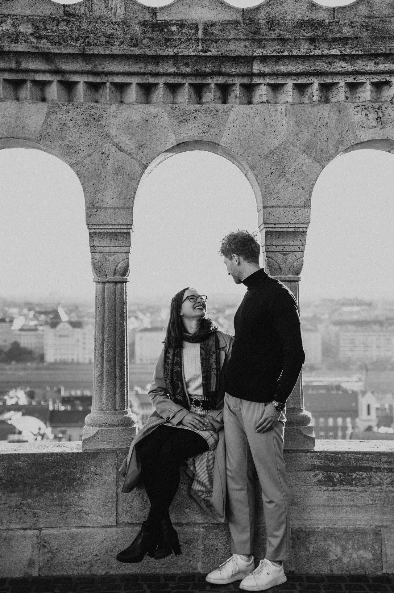 Black-and-white photograph of the woman sitting beside her partner in Fisherman’s Bastion’s stone arcade with the skyline beyond during a couple photoshoot in Budapest.