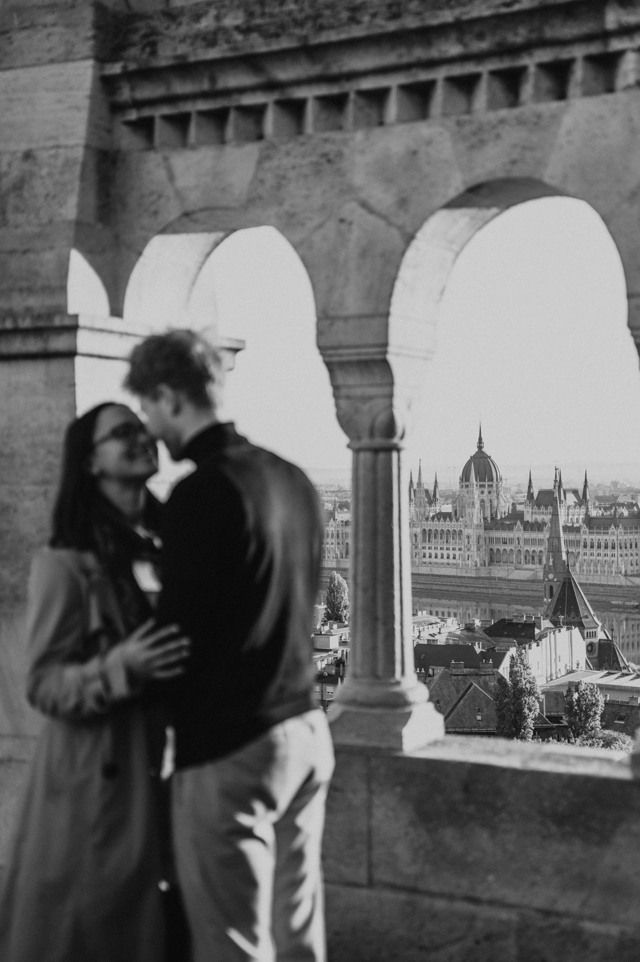Soft-focus black-and-white portrait of the couple embracing in Fisherman’s Bastion’s arcade with Parliament in the background during a couple photoshoot in Budapest.