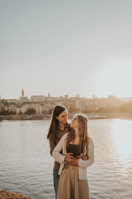 Budapest couple photoshoot beside the Danube in sunset light The couple embracing by the Danube during their Budapest photoshoot, with the skyline across the river and warm sunset light behind them.