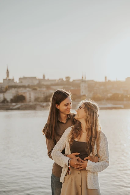 Couple photoshoot in Budapest overlooking the opposite Danube riverbank The couple smiling towards each other in an embrace during their Budapest photoshoot beside the Danube, with the opposite riverbank behind them.