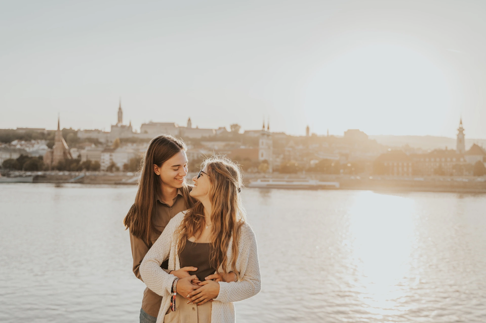 Budapest photoshoot with warm river light and the city skyline Wide portrait of the couple embracing during their Budapest photoshoot, with the Danube and a warmly lit skyline across the river.