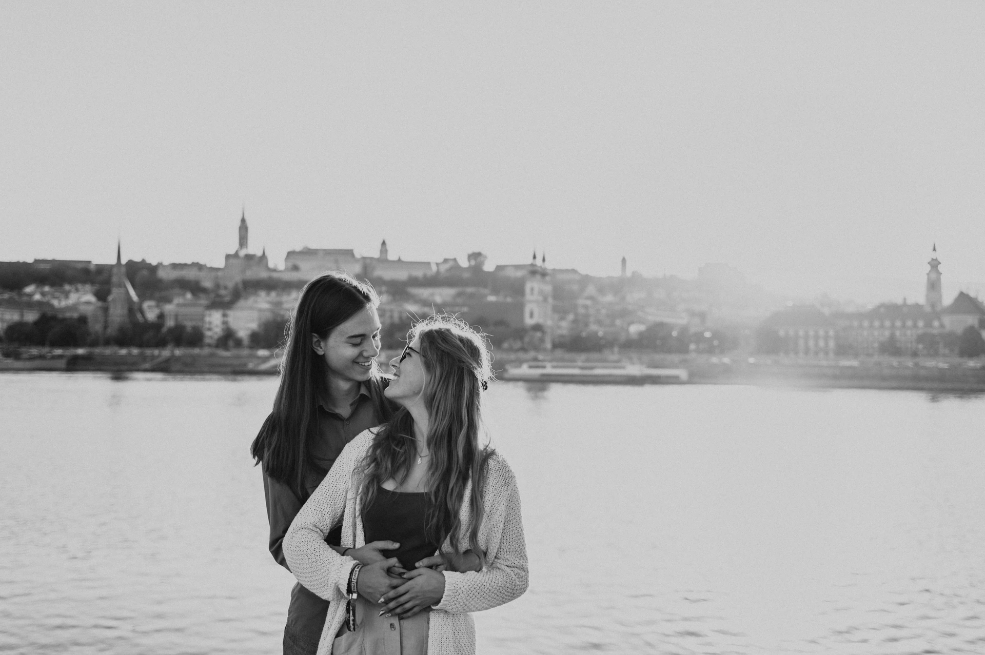 Black-and-white wide portrait of the couple embracing beside the Danube during their Budapest photoshoot, with the opposite skyline behind them.