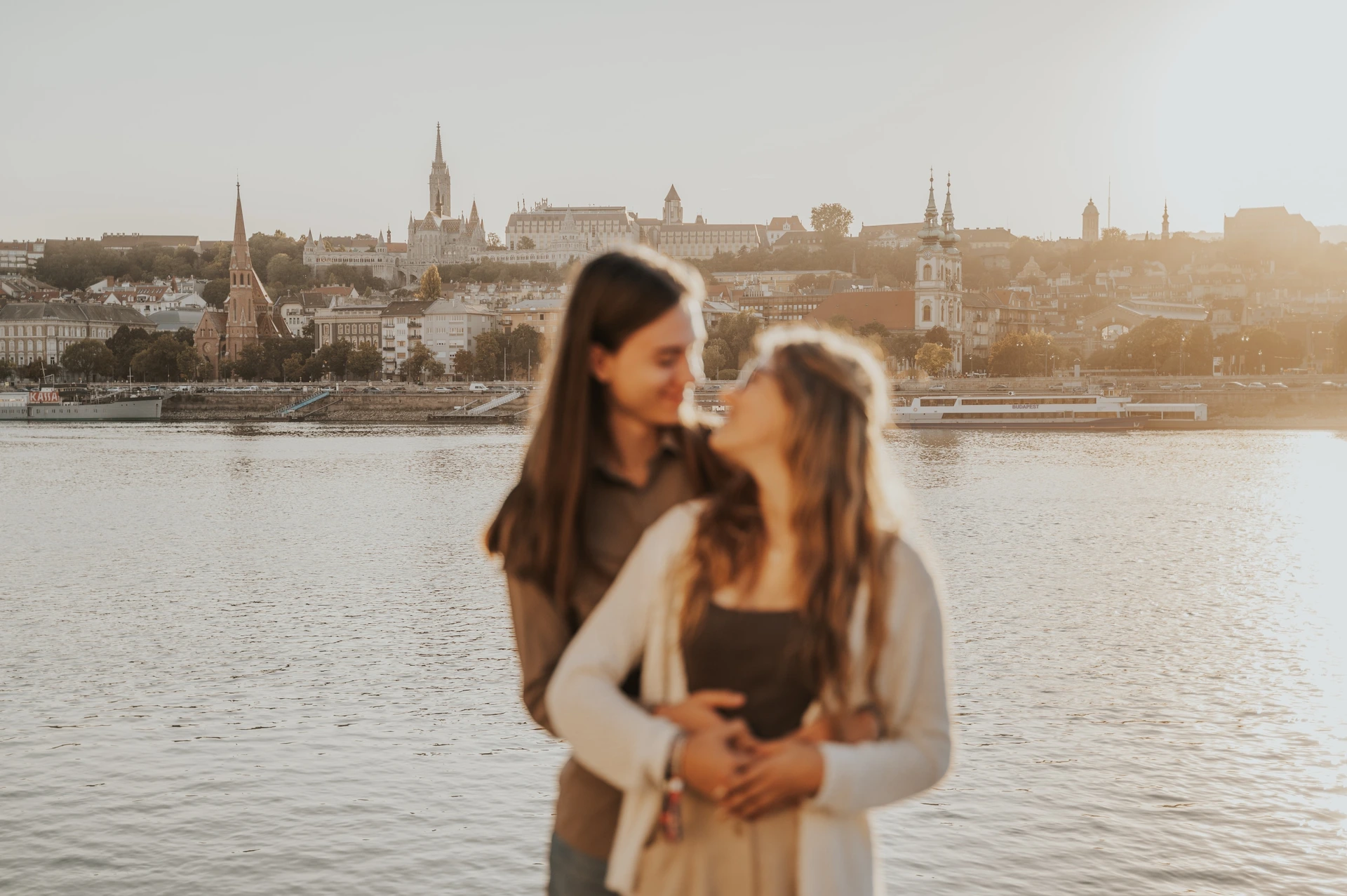 A closer wide portrait of the couple smiling at each other during their Budapest photoshoot beside the Danube in warm evening light.