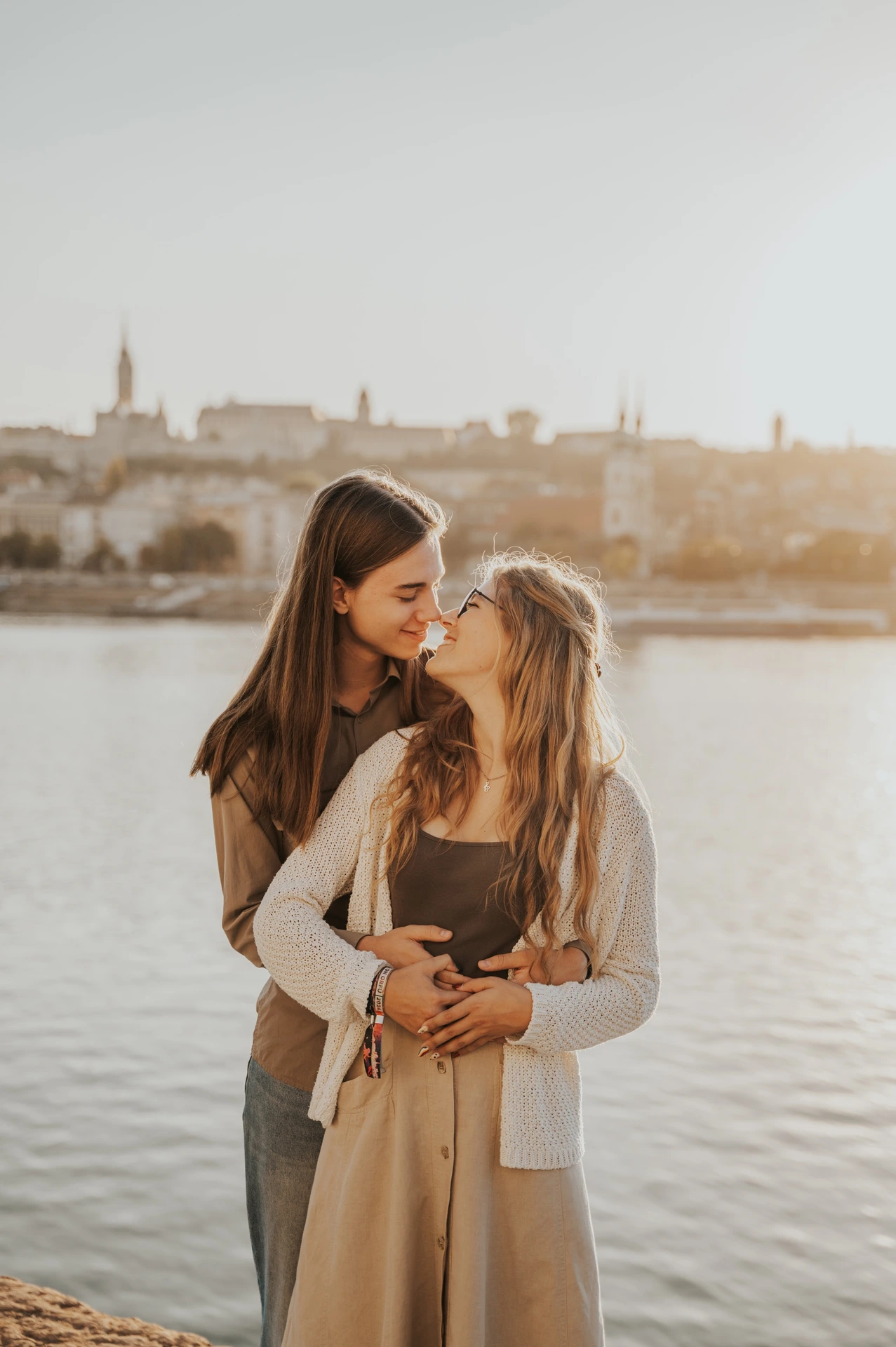 The couple looking towards each other while embracing during their Budapest photoshoot by the river, with soft sunlight on their hair.