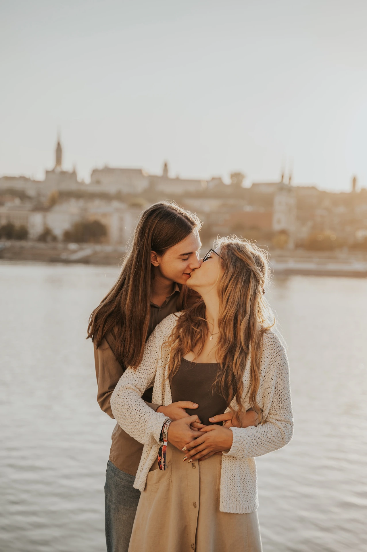 The couple leaning close together during their Budapest photoshoot beside the Danube, with the city across the water in the backdrop.