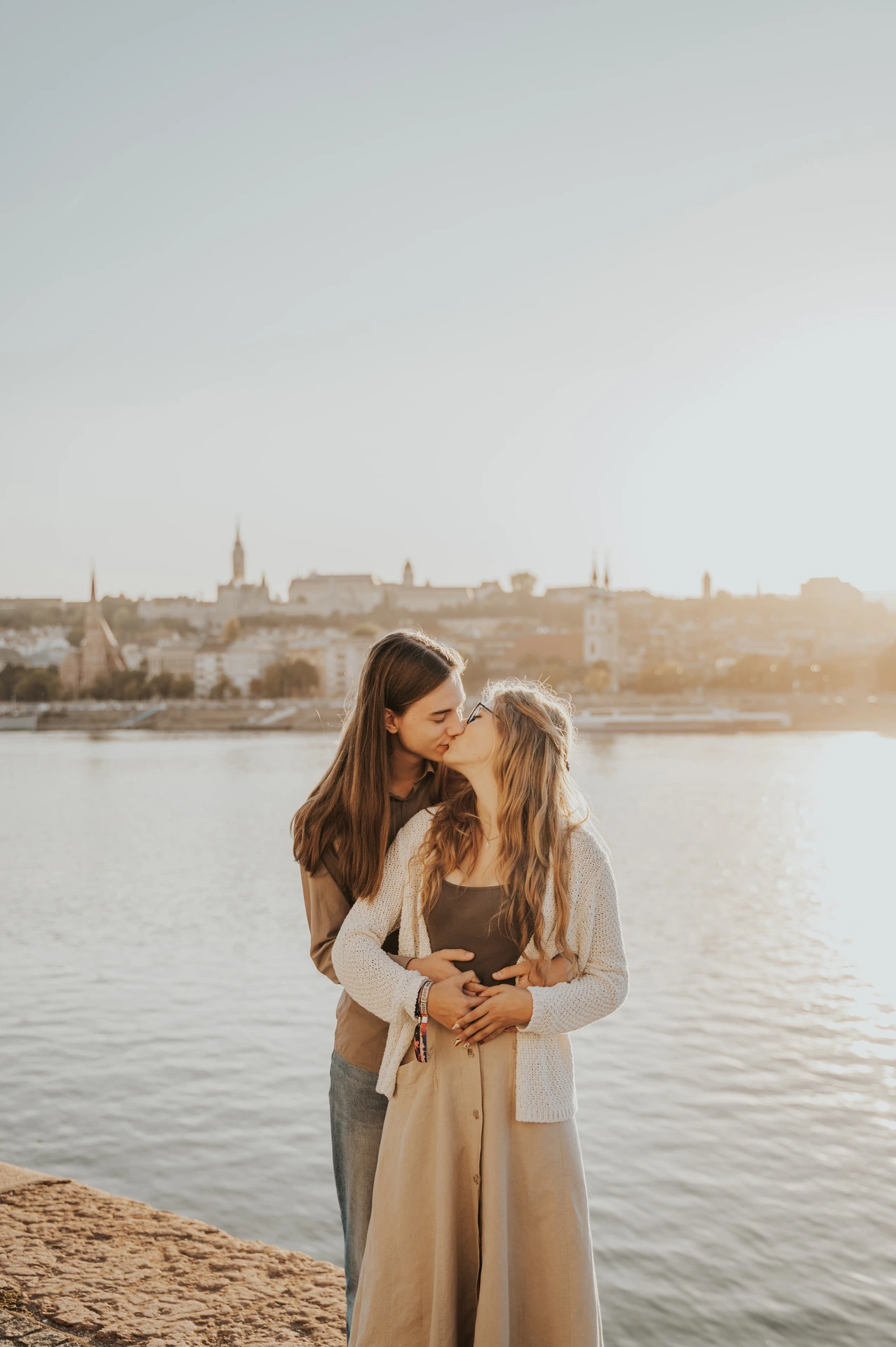 A wide vertical view of the couple embracing by the Danube during their Budapest photoshoot, with the evening sky above the water.