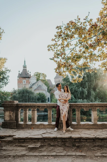 An embrace by the stone balustrade at a Budapest couple photoshoot The couple embracing beside a stone balustrade with Vajdahunyad Castle’s turrets and golden leaves in the background during their couple photoshoot in Budapest.